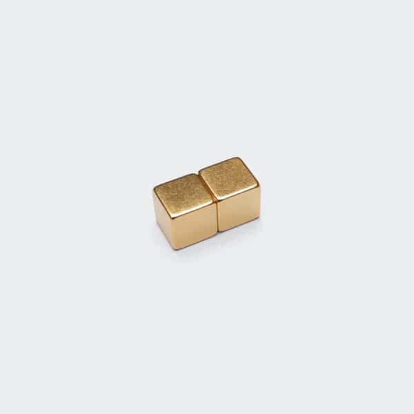 Stack of two tiny neodymium cube magnets with shiny gold plated surface treatment, laid flat on a white desktop.