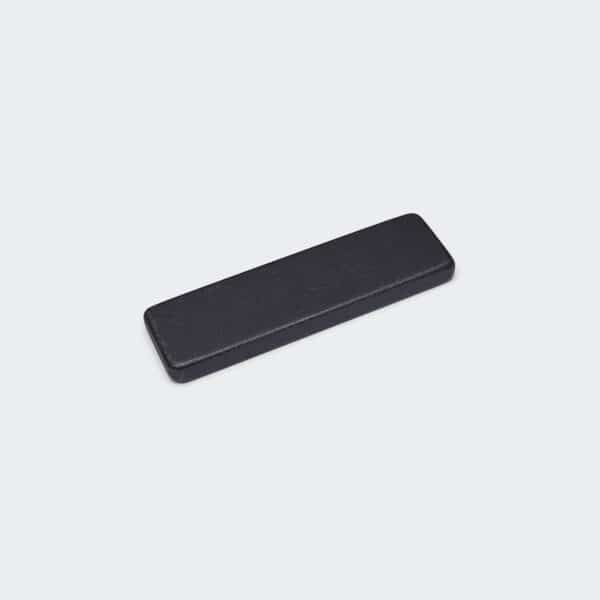 A thin rectangular strong plate magnet with black PTFE coating, laid flat on a white desktop.